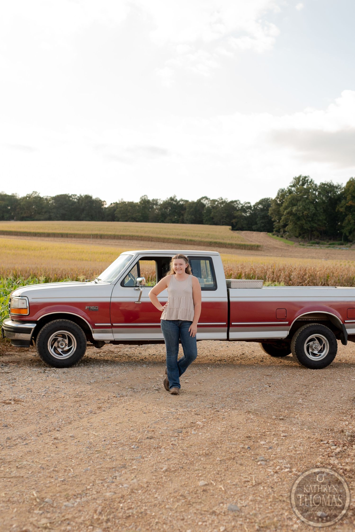 A red and white vintage Ford pickup truck parked on a rural dirt road next to a cornfield.
