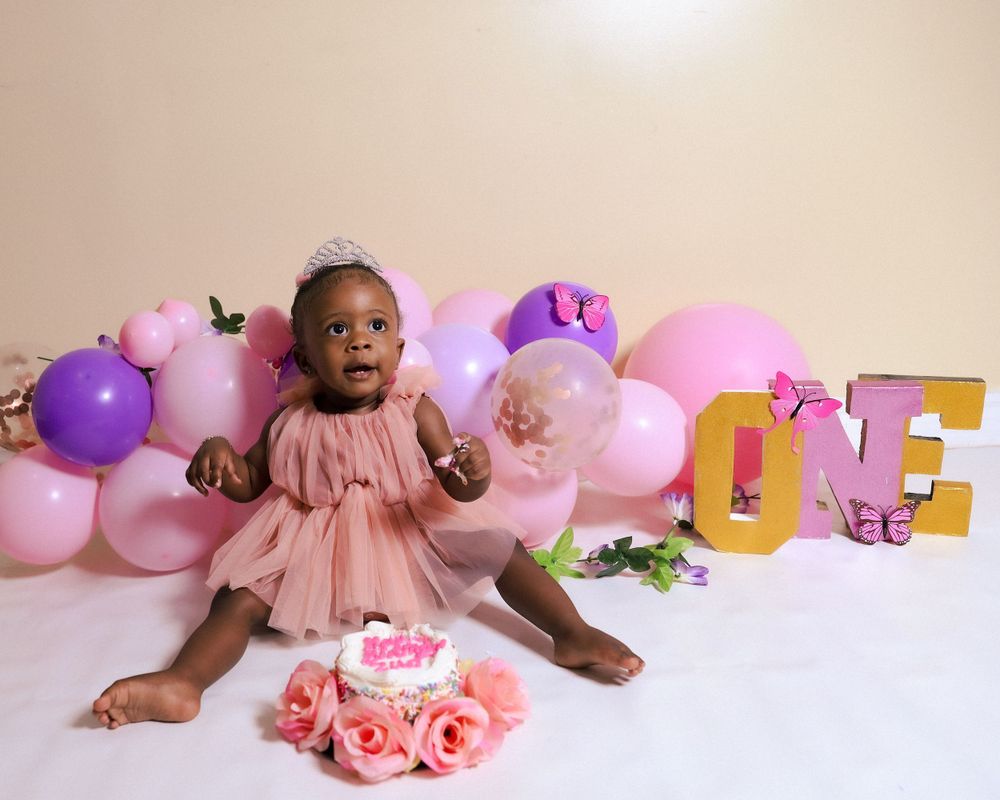 First birthday celebration setup with pink dress, balloons and cake against pink and purple decor.