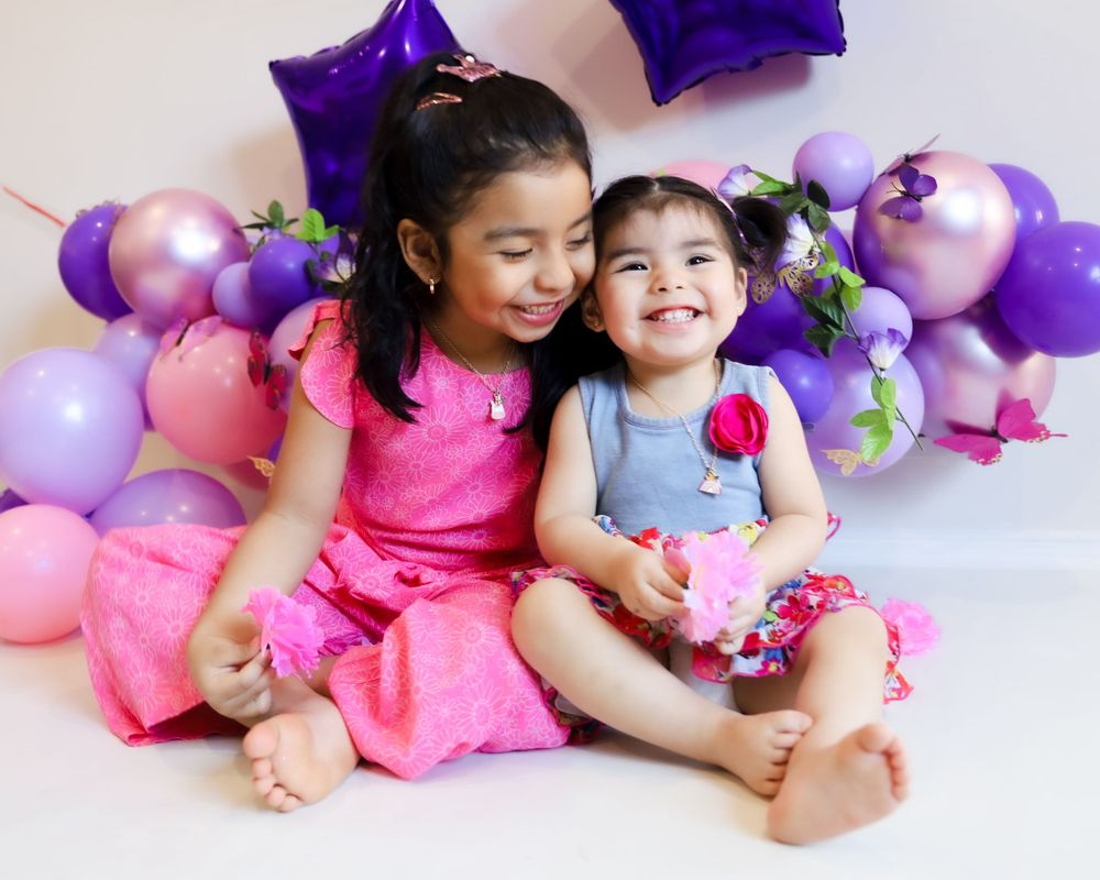 Two children sitting together against pink and purple balloon decorations.