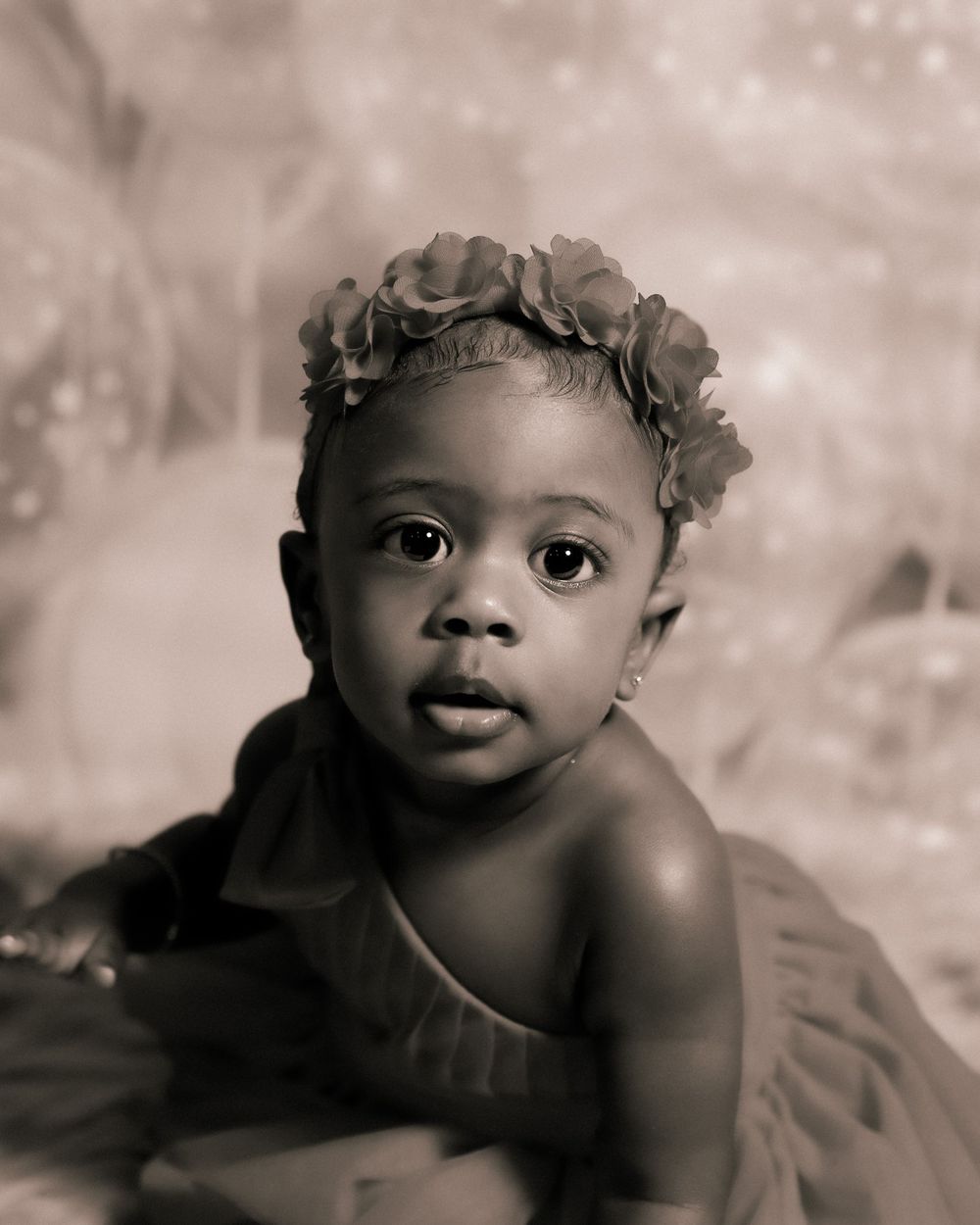 A vintage black and white portrait of a baby wearing floral crown looking up with curious eyes.