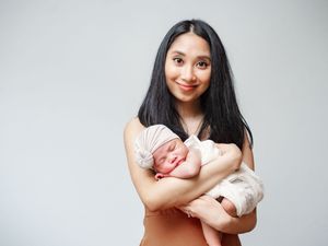 Young parent in white top holds sleeping newborn baby against gray background in tender embrace.