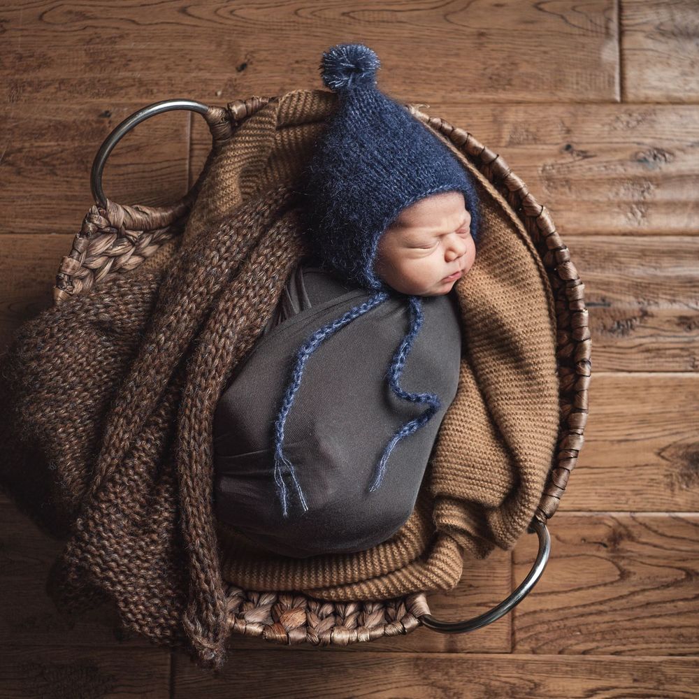 Sleeping baby wrapped in cozy knit blankets and wearing blue winter hat rests in rustic wooden basket.