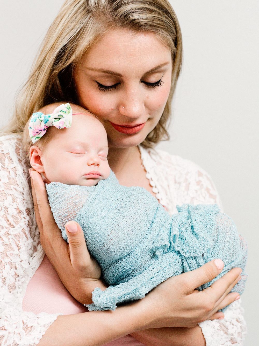 Sweet tender moment as parent in lace gently holds sleeping newborn swaddled in soft blue wrap with floral headband.