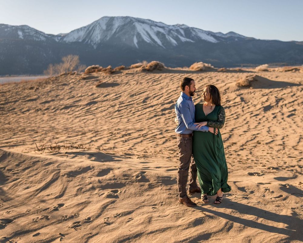 A couple embraces on sandy dunes with snowy mountain peaks rising dramatically in the background.
