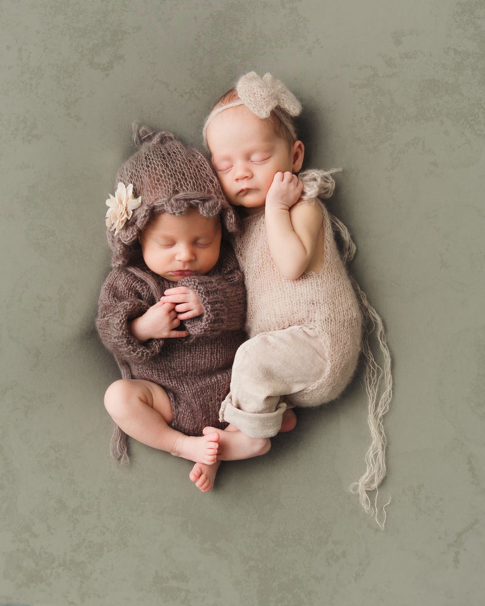Two sleeping infants in cozy knit outfits pose together on textured grey background for newborn photography.