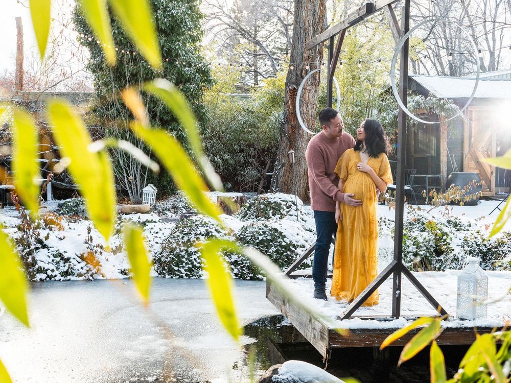 A couple stands on a snowy wooden bridge in a garden setting, with yellow foliage framing the winter scene.