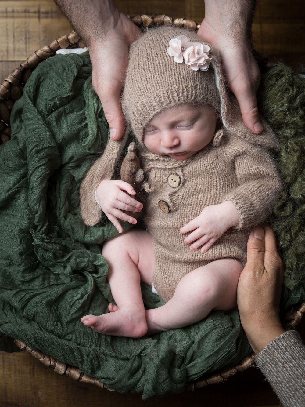Sleeping newborn wearing cozy knitted beige romper and hat rests on dark green textured blanket in basket.
