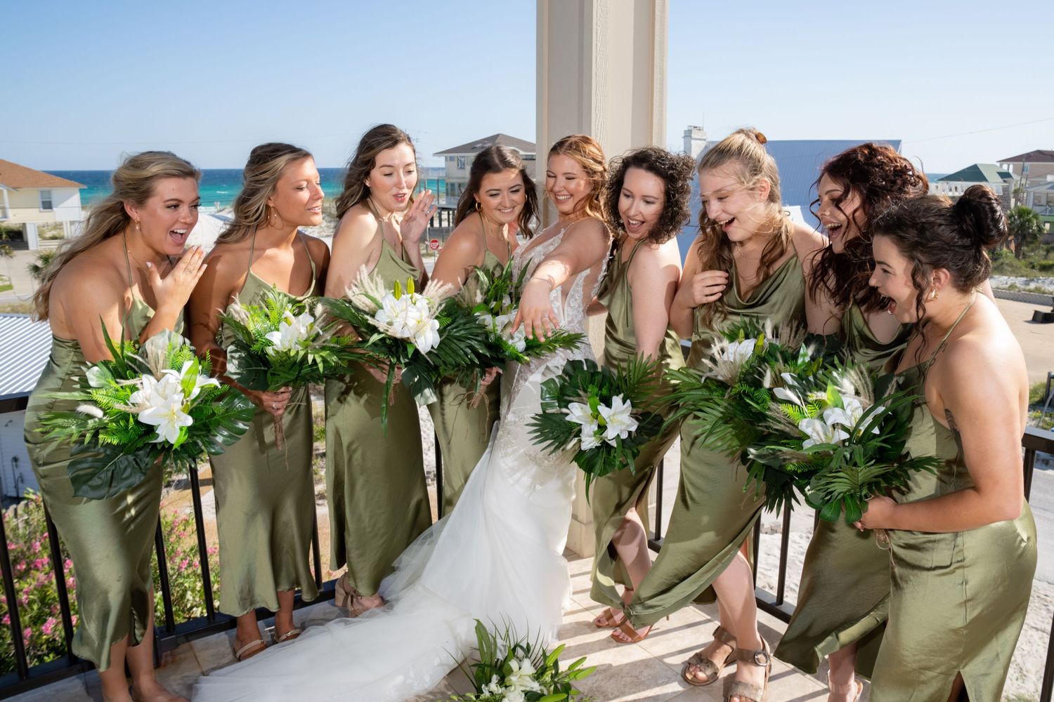 Bridesmaids in sage green dresses gather together holding white floral bouquets on a sunny balcony.