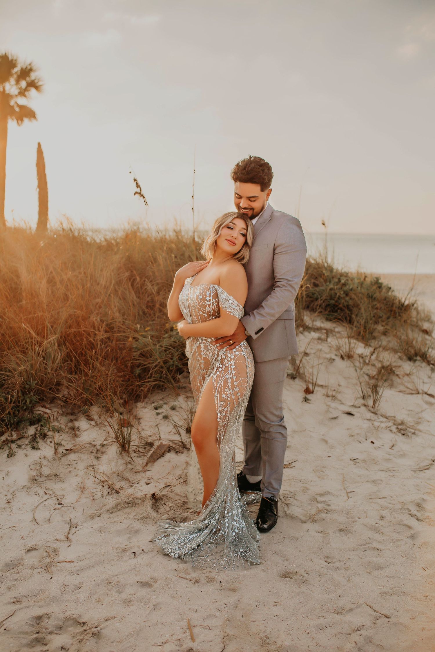 Couple embraces on sandy beach at sunset wearing formal attire.