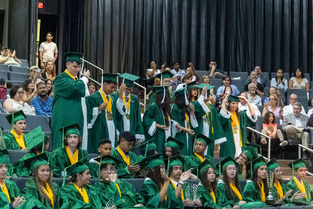 Graduates in emerald green caps and gowns sit together during commencement ceremony.