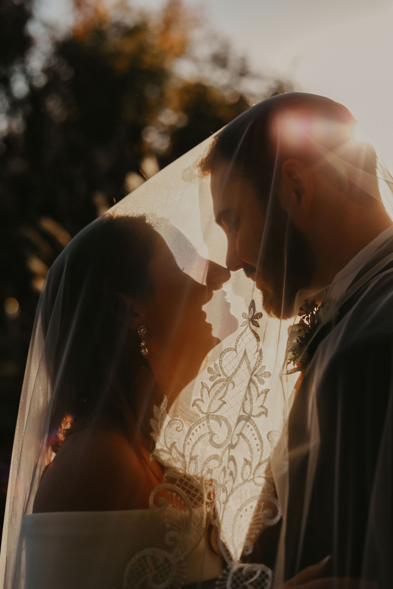 Romantic silhouette of newlyweds sharing an intimate moment under a sheer veil at sunset.