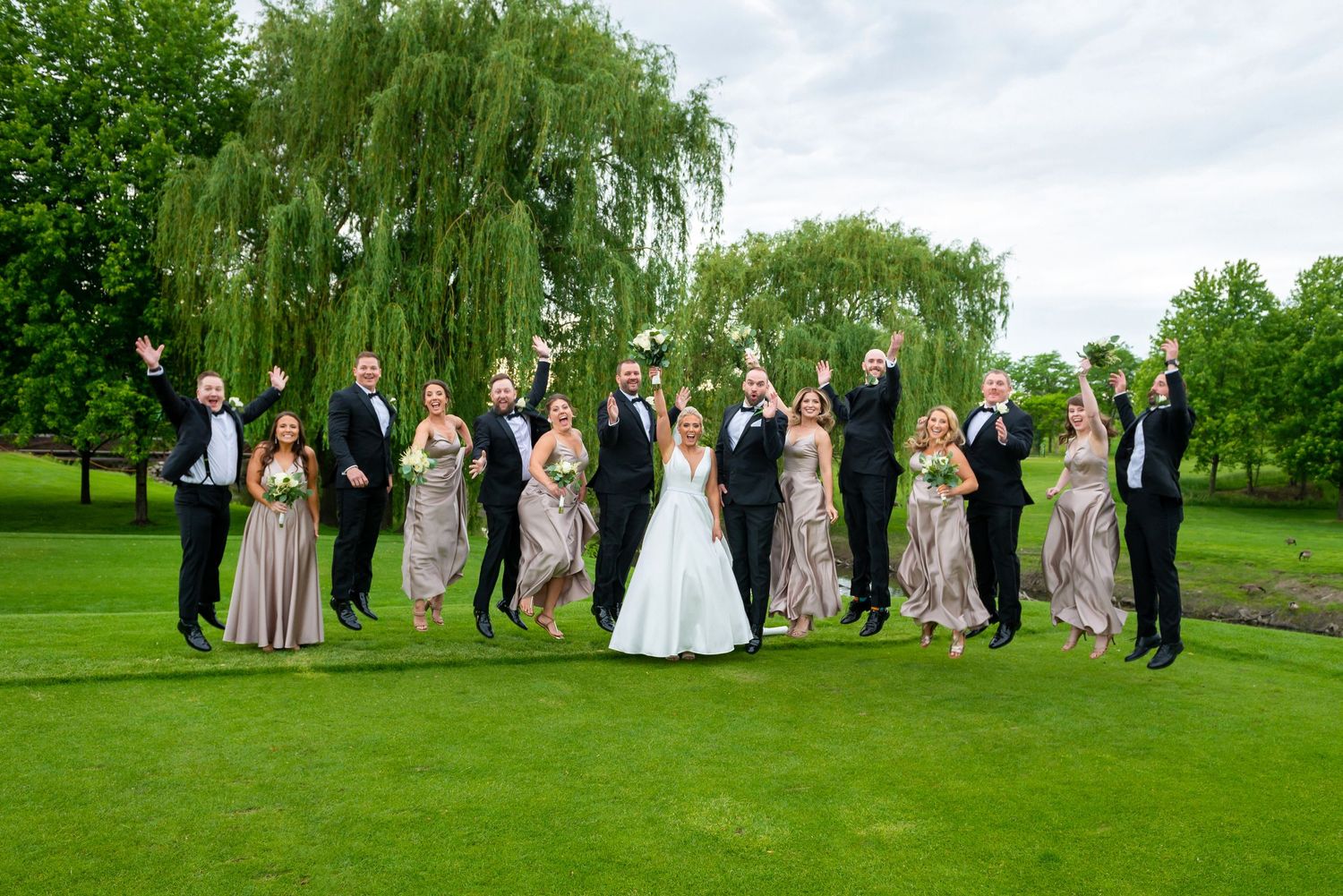 Wedding party lined up jumping in the air on grass with trees in the background.