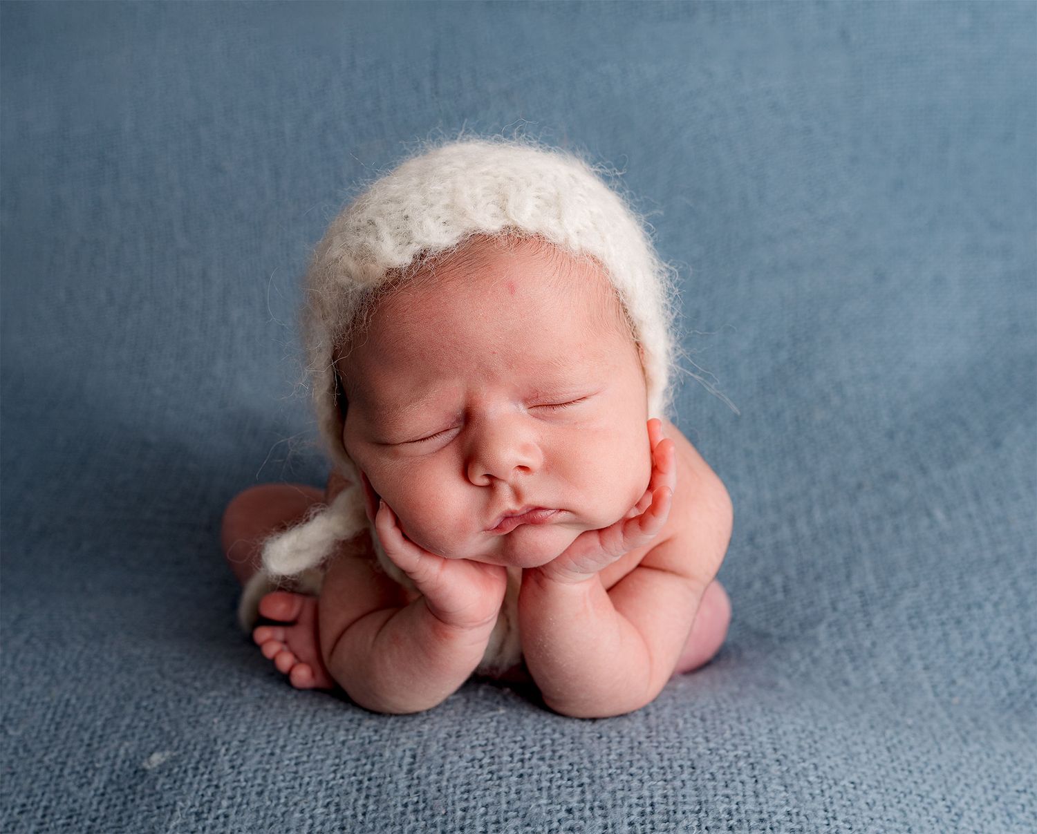 A froggy composition of a sweet sleeping newborn wearing white bonnet rests on blue backdrop.