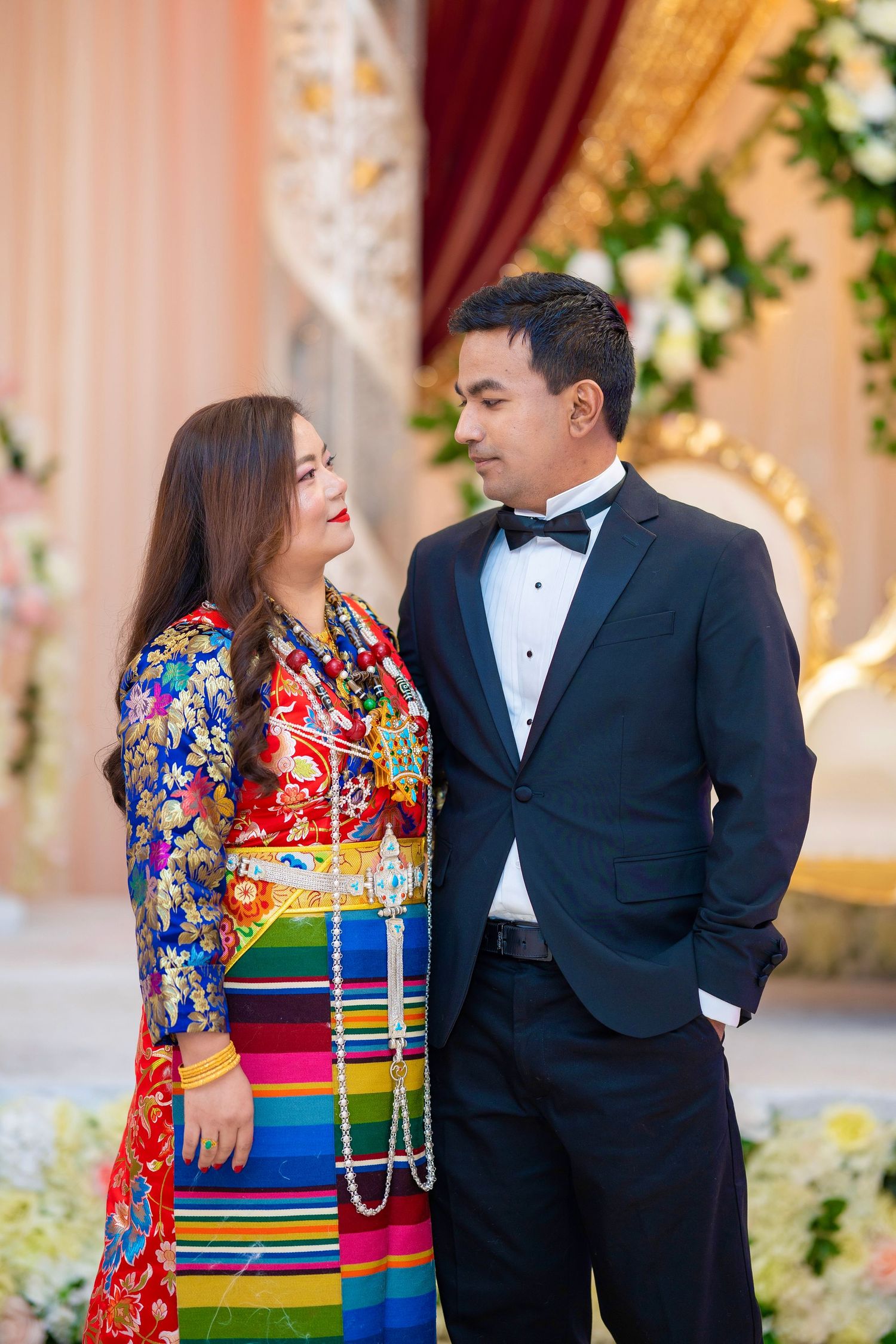 Elegant couple in traditional Nepali dress and formal tuxedo at decorated venue.