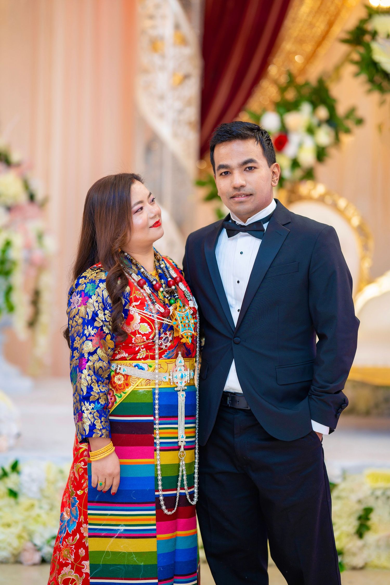A couple poses in formal attire, with the woman wearing a vibrant traditional dress featuring colorful stripes and patterns.