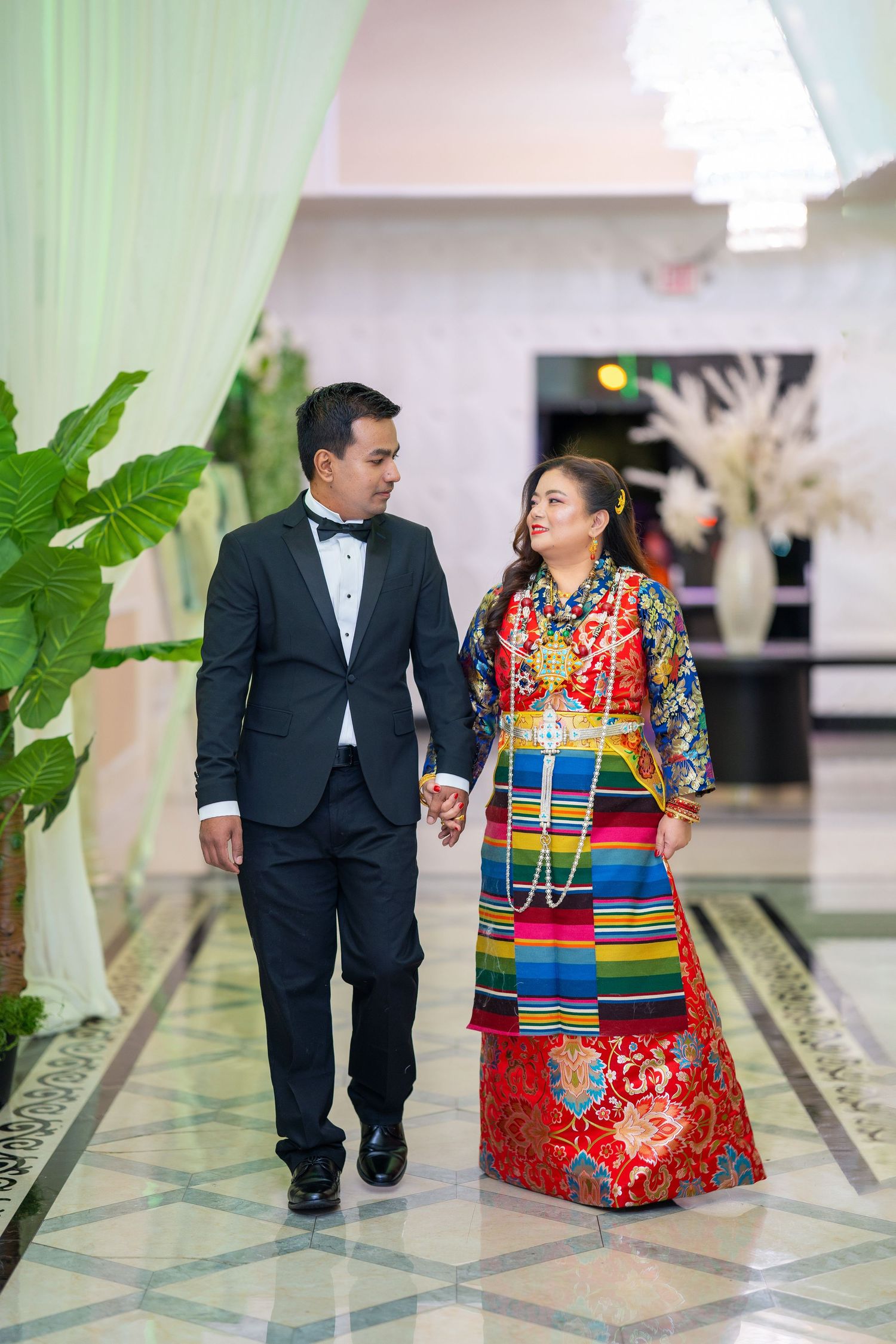 A couple walks together in an elegant venue, dressed in contrasting formal and traditional outfits.