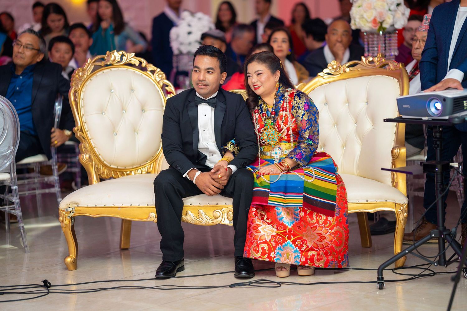 A couple sits on elegant cream-colored throne chairs during a formal celebration.