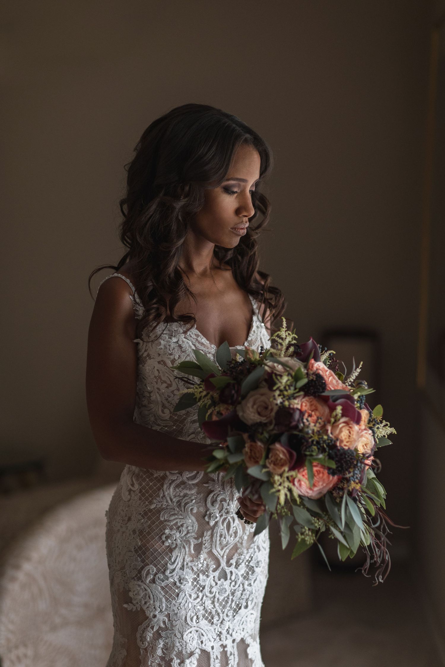 A bride in an elegant lace dress holds a vibrant pink and green bouquet against a dark background.