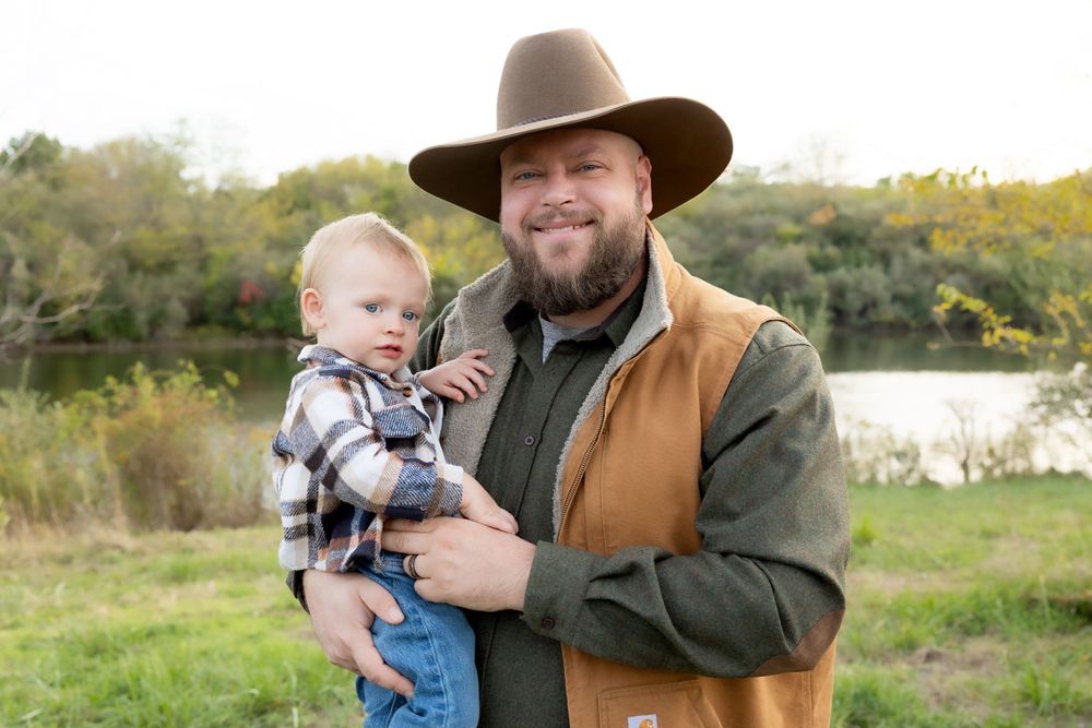 Adult wearing brown hat and vest holds small child outdoors by lake.