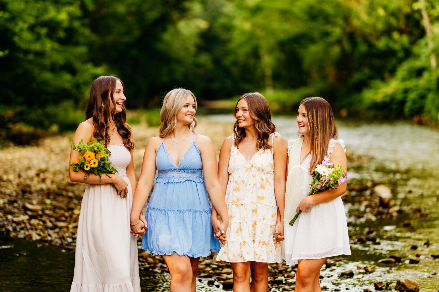 Four bridesmaids in pastel dresses laugh and hold hands by a peaceful creek surrounded by lush green trees.