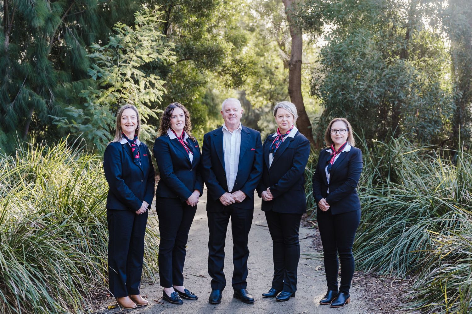 Professional team in business attire standing together on outdoor path surrounded by greenery.