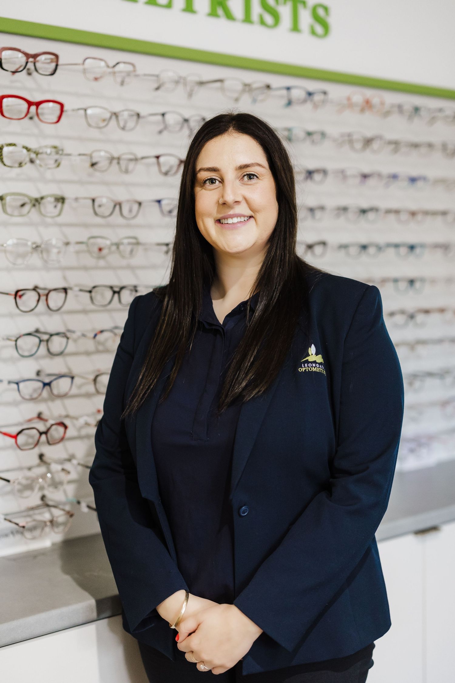 Staff member in navy blazer standing inside optometry store with eyewear display wall.