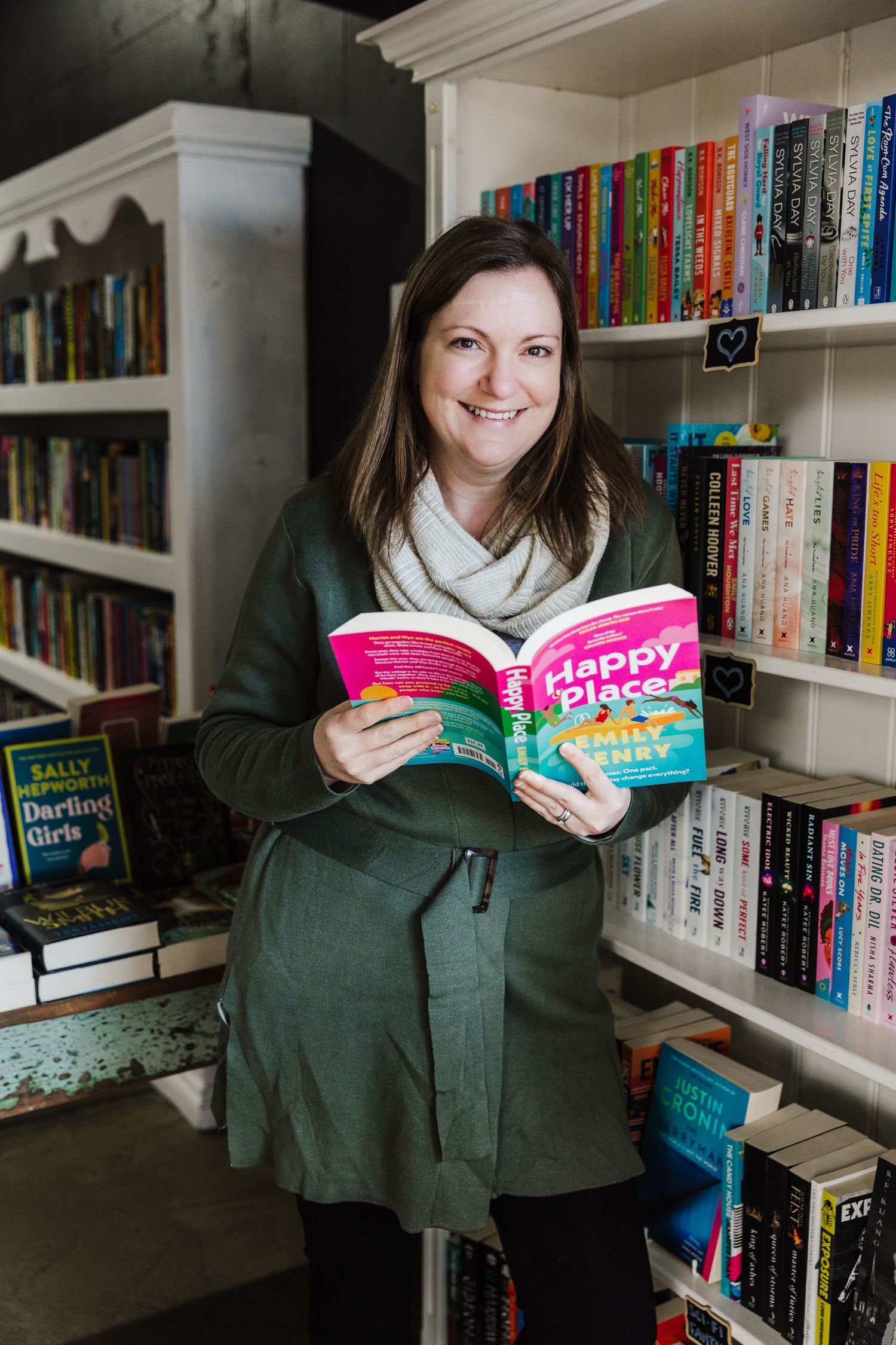 A book enthusiast browses a colorful library shelf while reading a bright pink book.