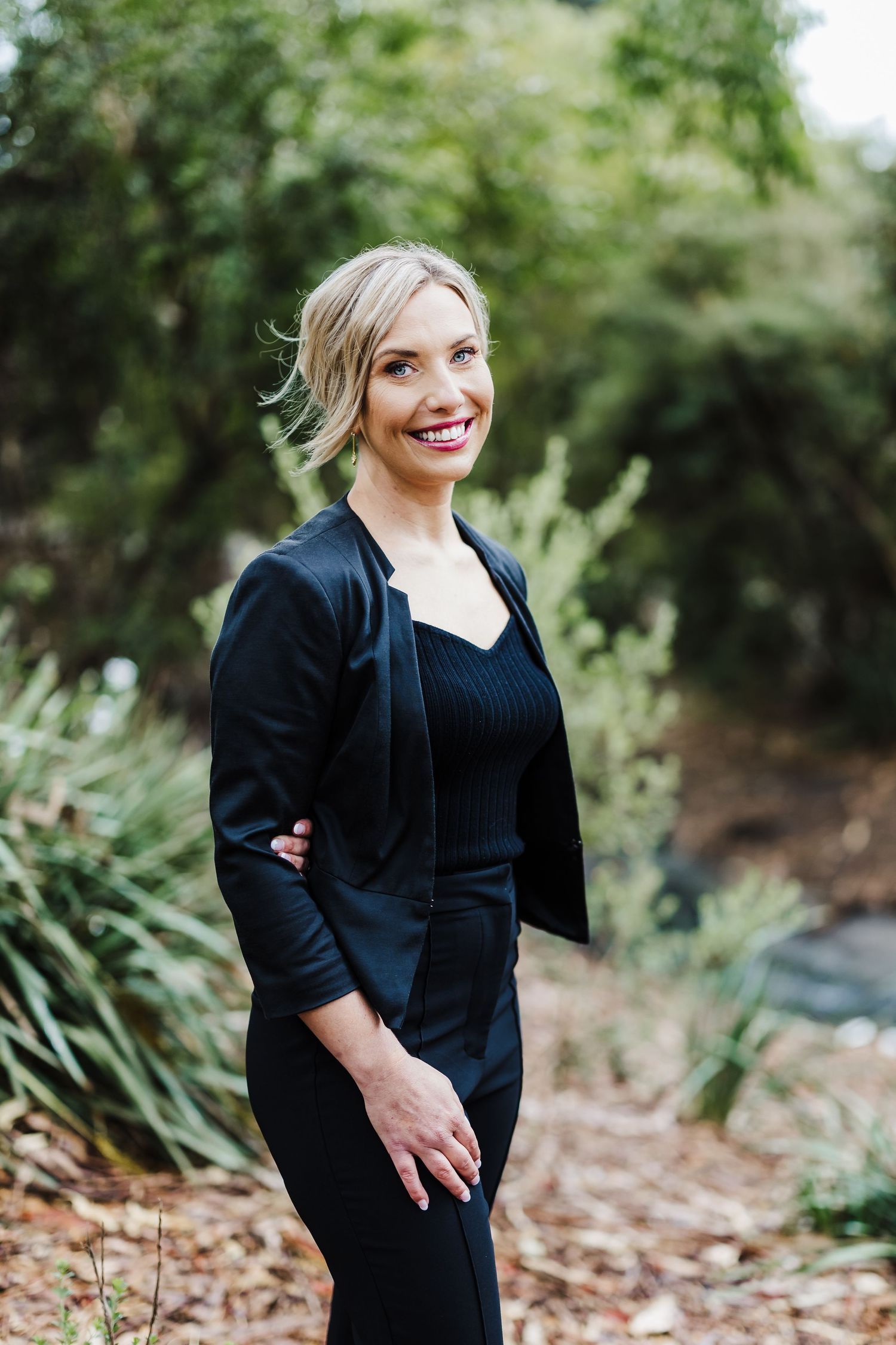 Professional in black outfit standing outdoors with natural garden background.