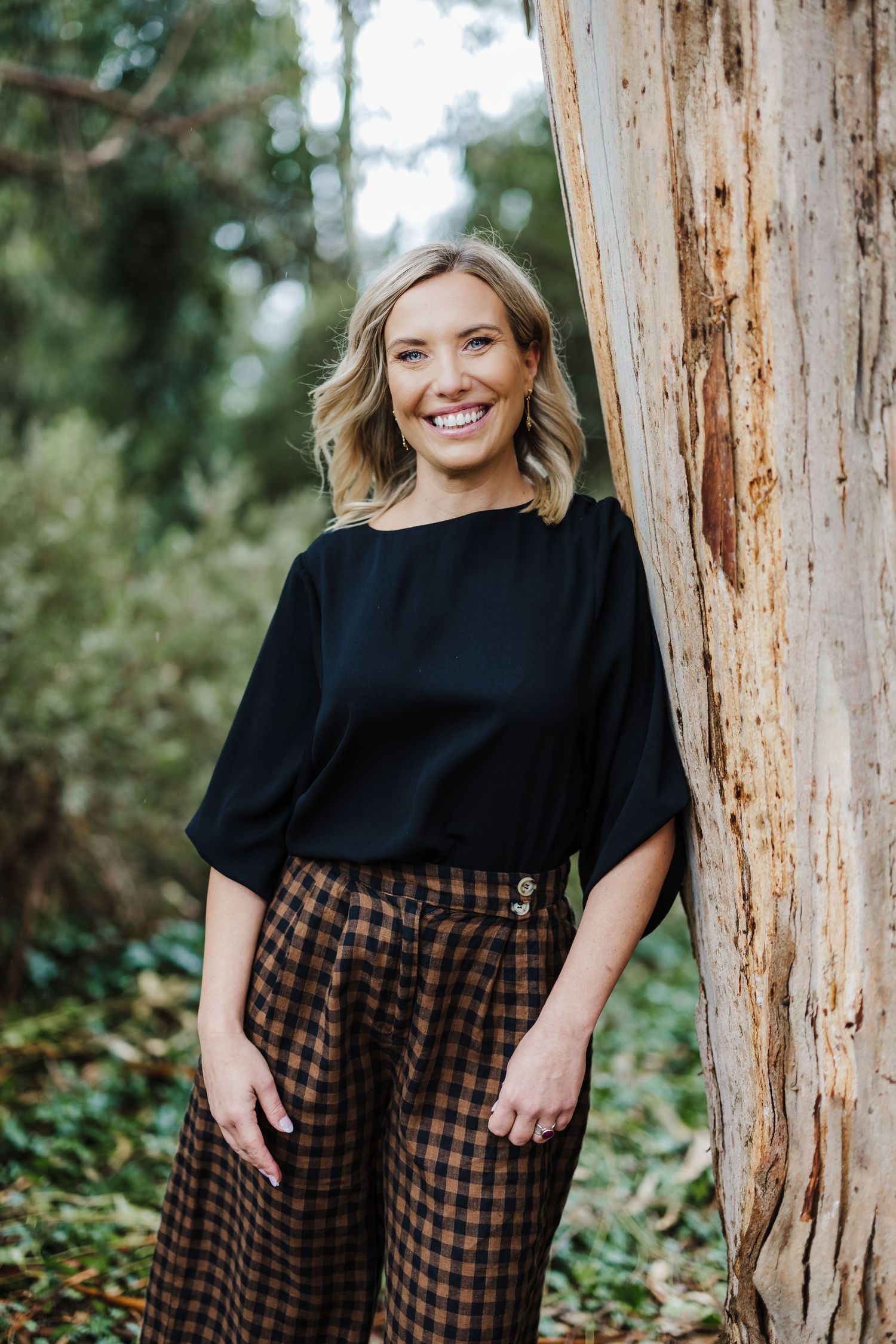 Professional in black top and plaid pants standing next to eucalyptus tree.