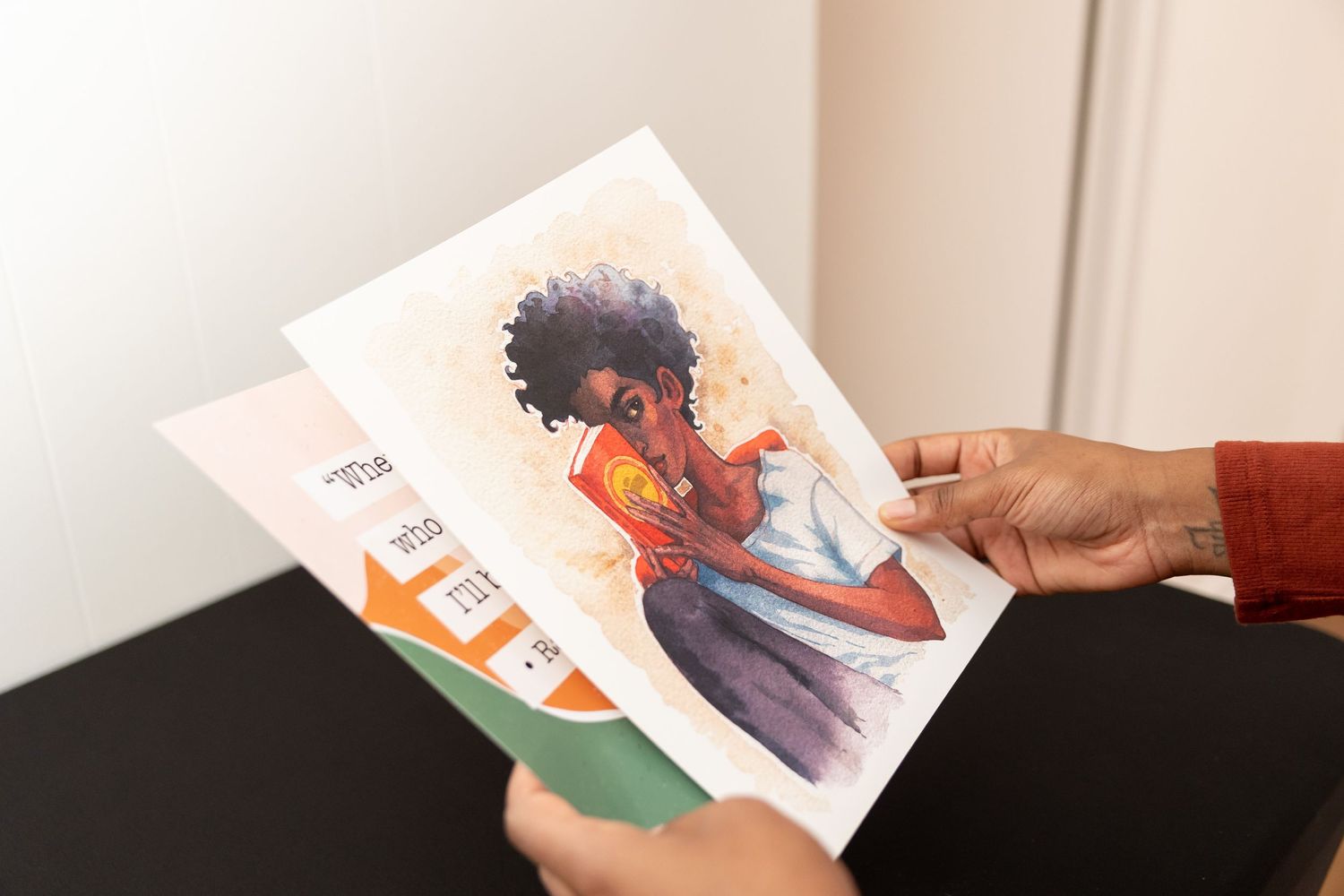 Hands holding a printed illustration of a Black woman with natural hair in watercolor style.