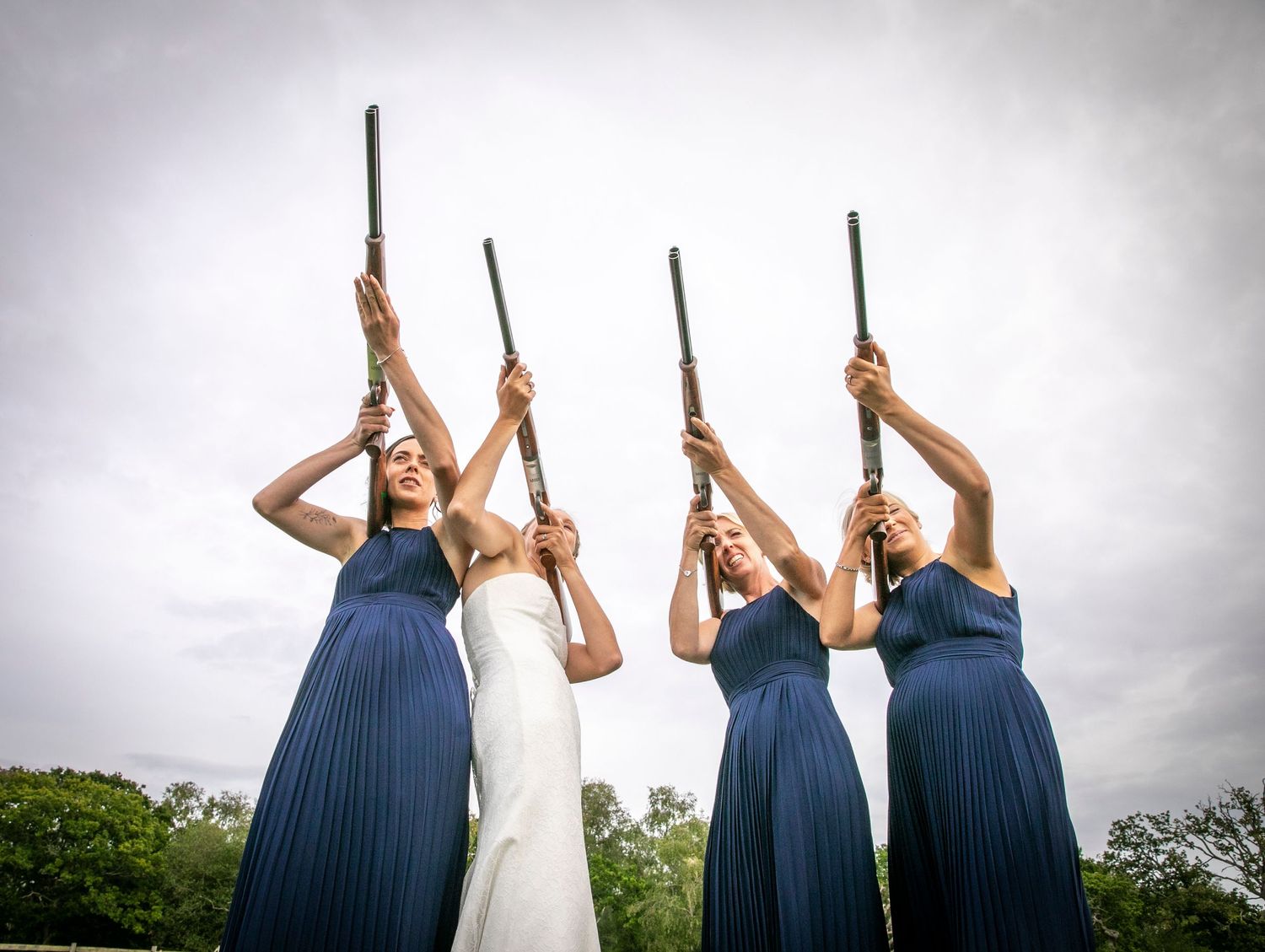 Wedding party posing with clay shooting rifles against cloudy sky.