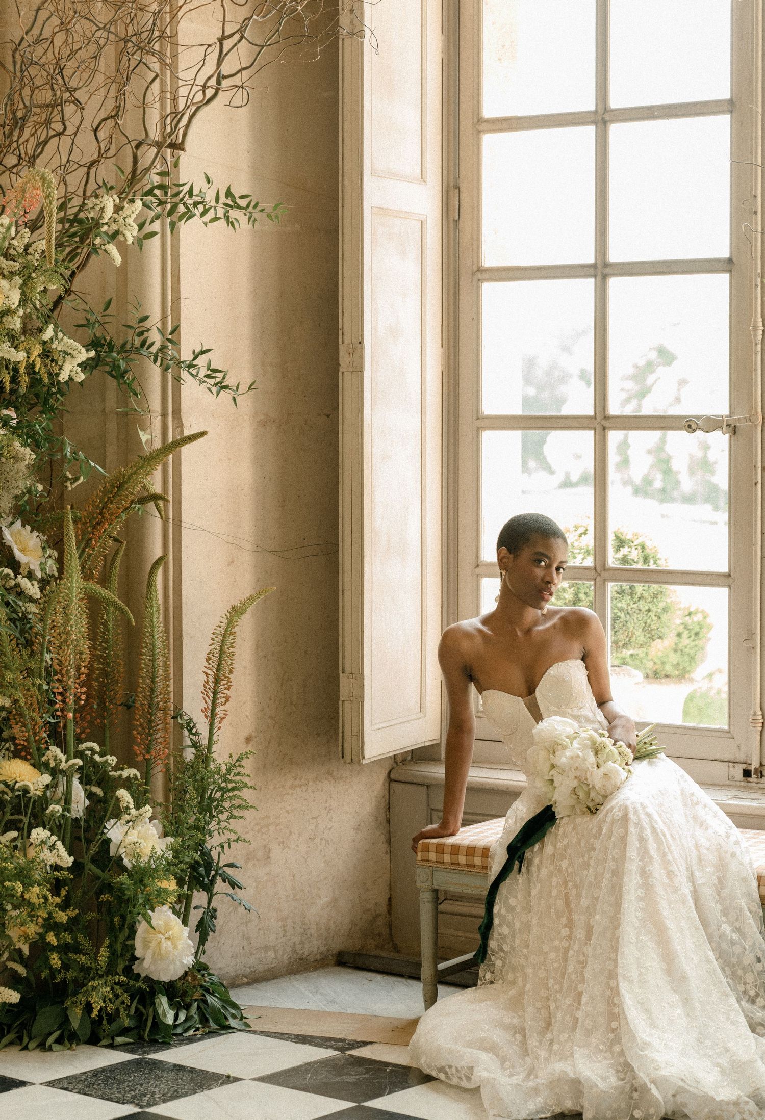 Romantic bridal portrait by tall window with natural light and floral arrangements.