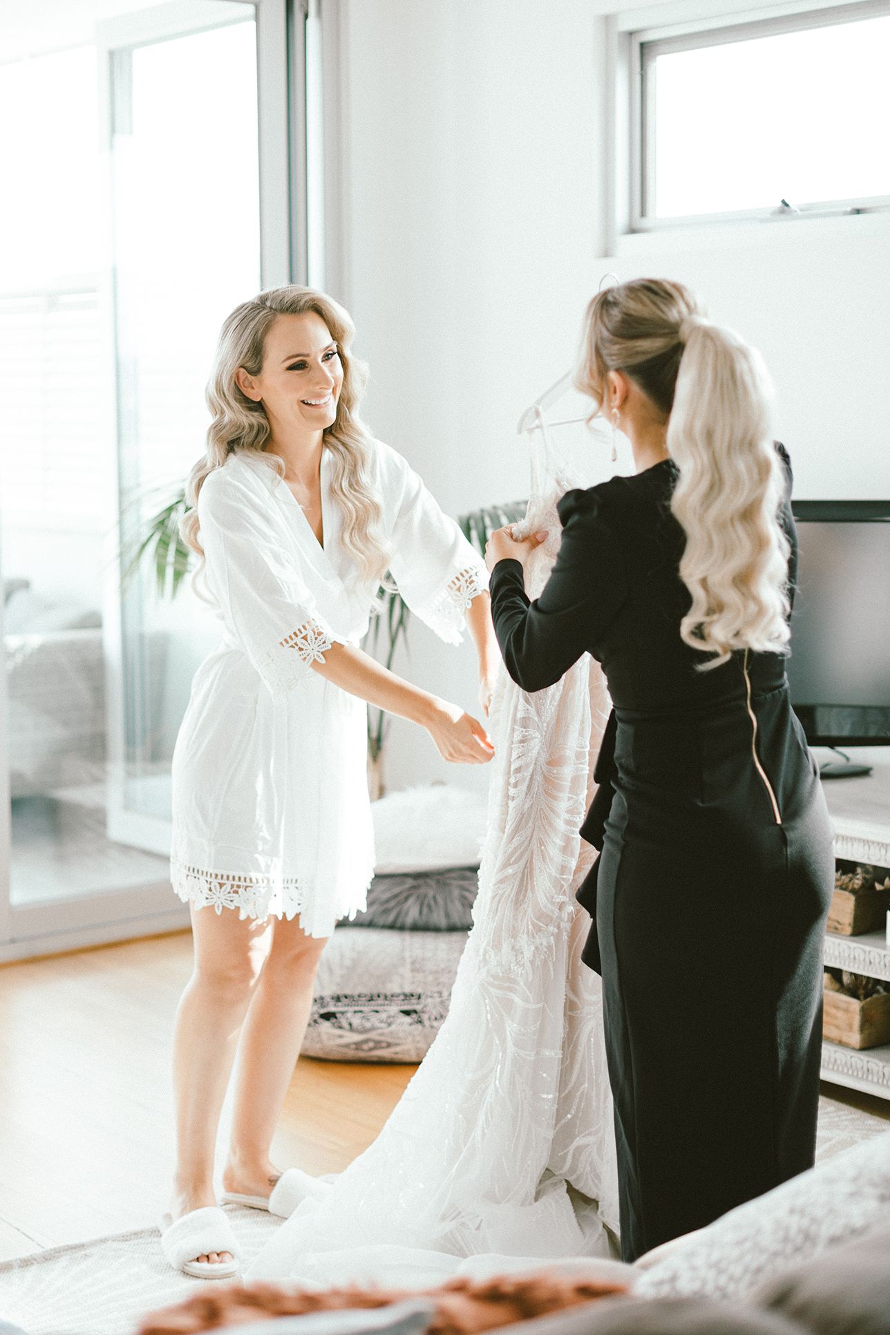 Two people prepare a wedding dress in a bright modern room with natural lighting.
