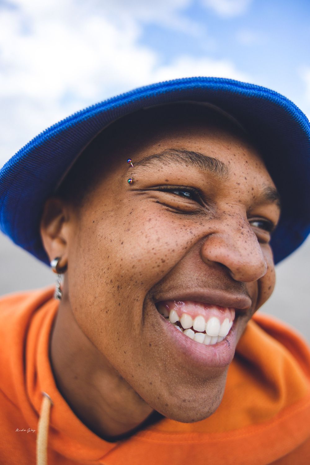 Close up portrait of someone wearing an orange hoodie and blue cap smiling brightly against a blue sky background.