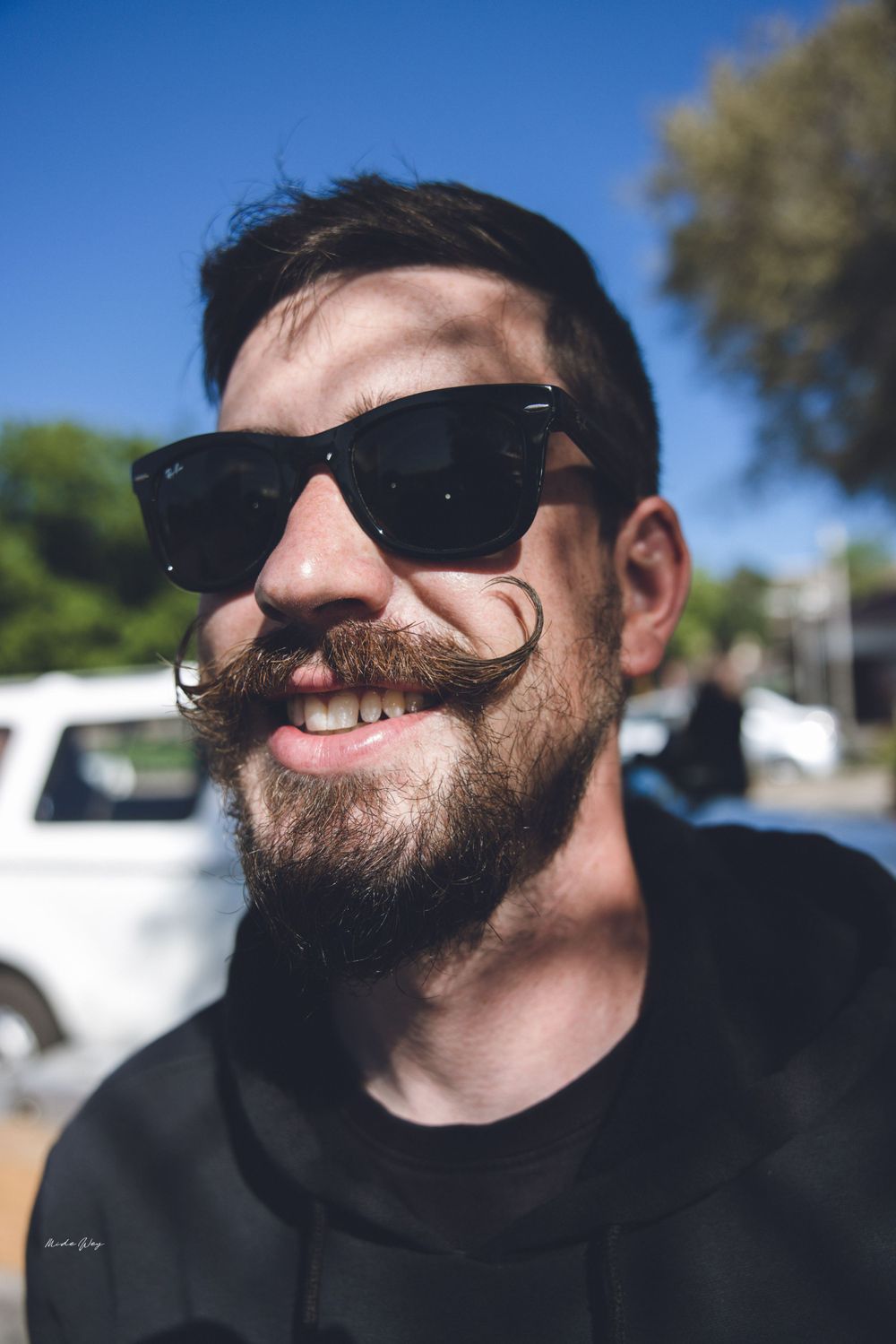Portrait of someone with a curled mustache wearing sunglasses and smiling outdoors.
