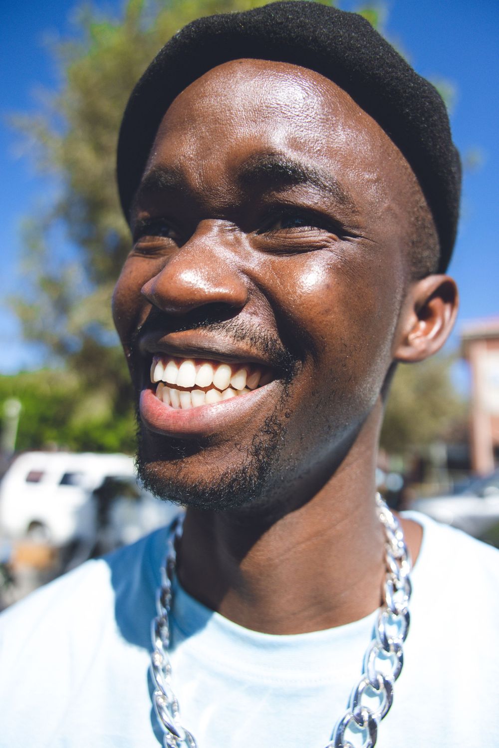 Portrait of someone wearing a beanie and chain necklace smiling broadly against a sunny outdoor background.