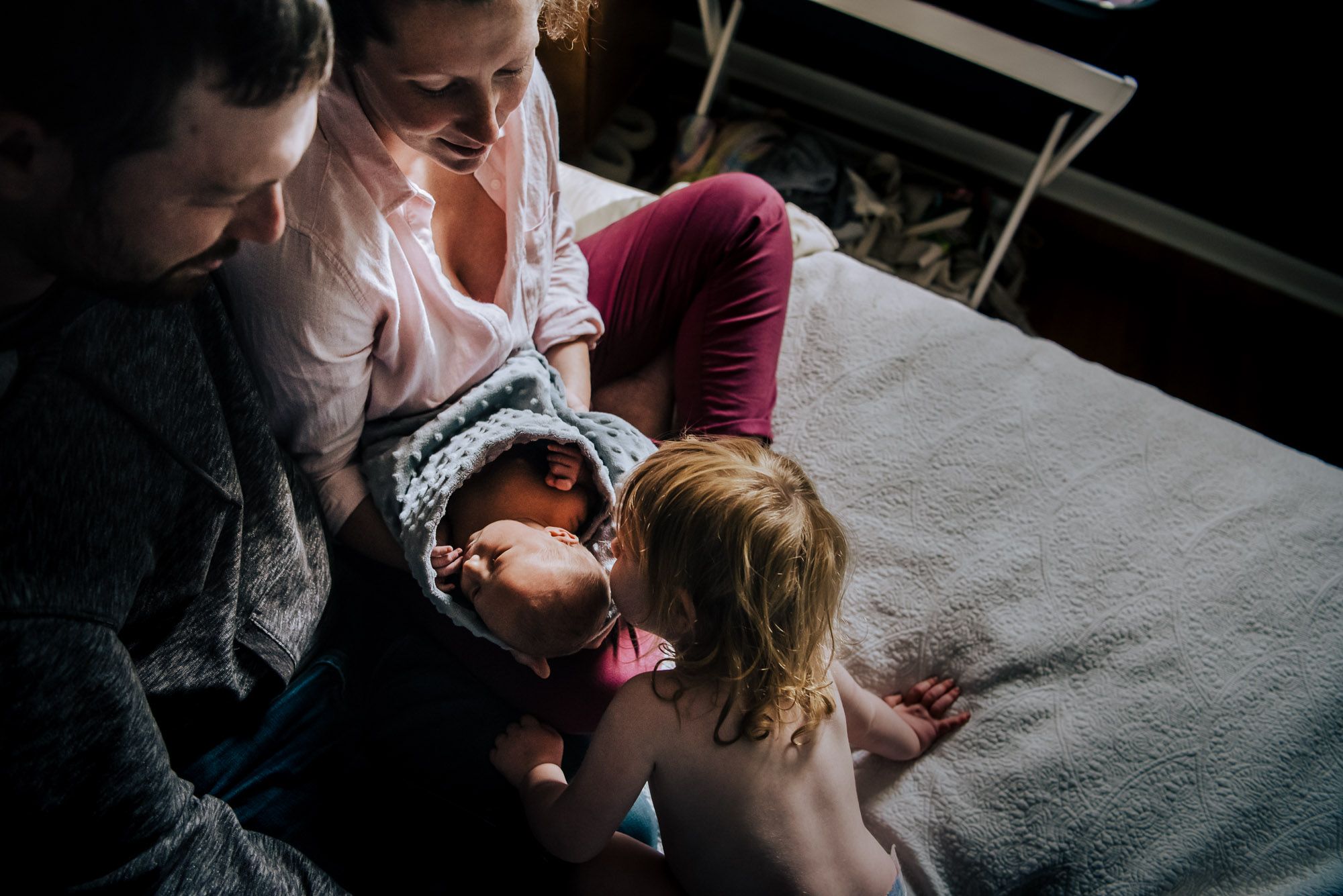 A newborn is held by her parents as they sit on their bed and her older toddler sister leans over to kiss her head.