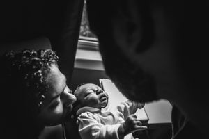 A father passes his newborn to his wife who is sitting down and kissing the baby's head.