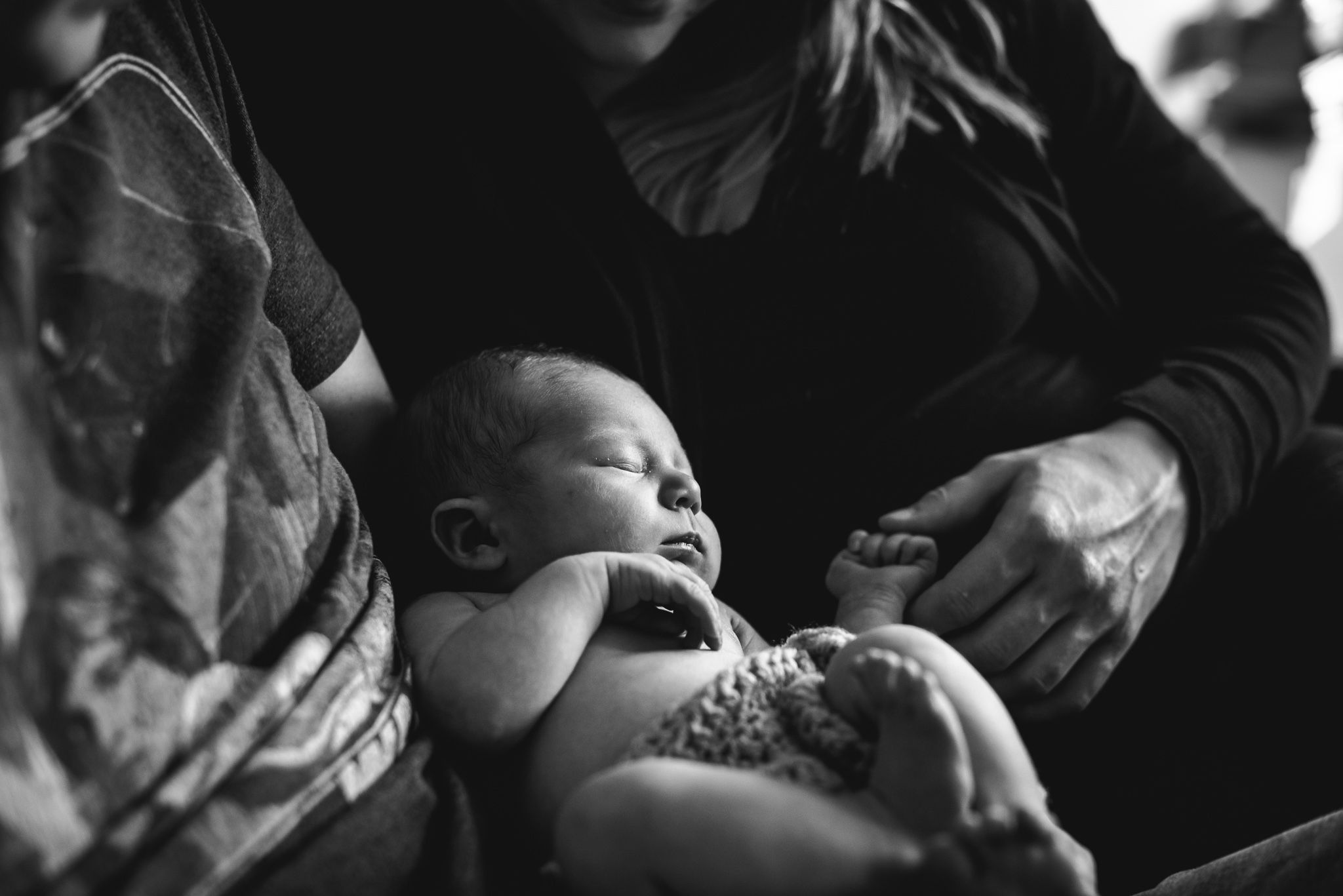 A newborn lays in her father's lap with her mother touching her little fist as she sleeps.