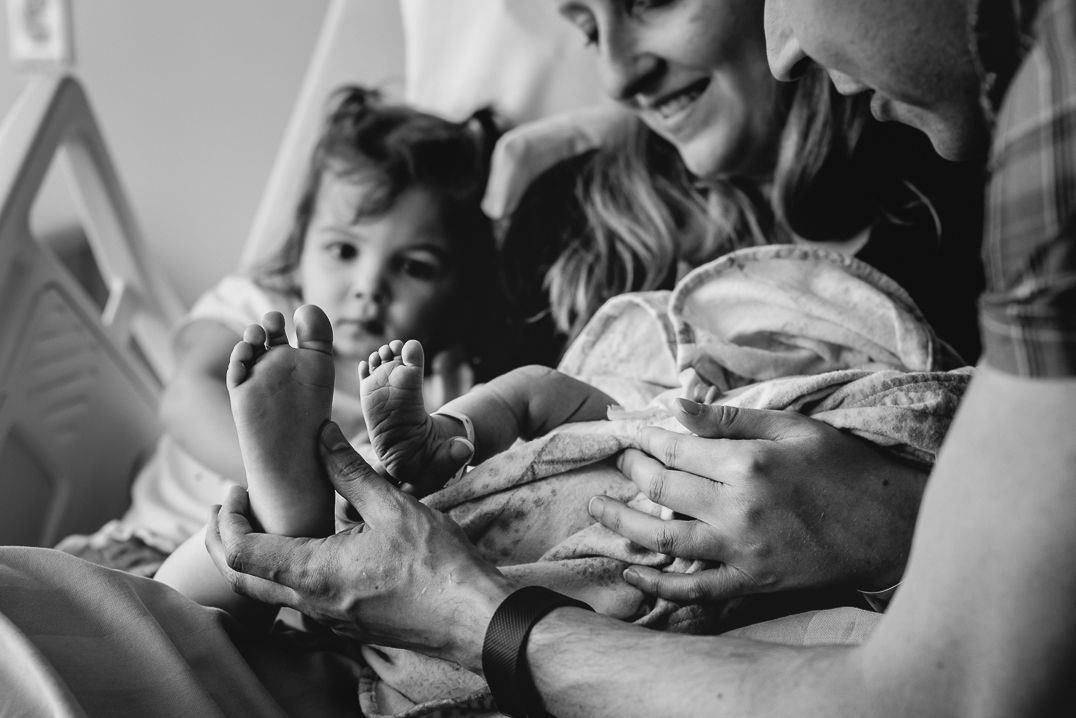 A family is snuggled up in a hospital bed with their newborn and holding her foot next to her older sister's foot.