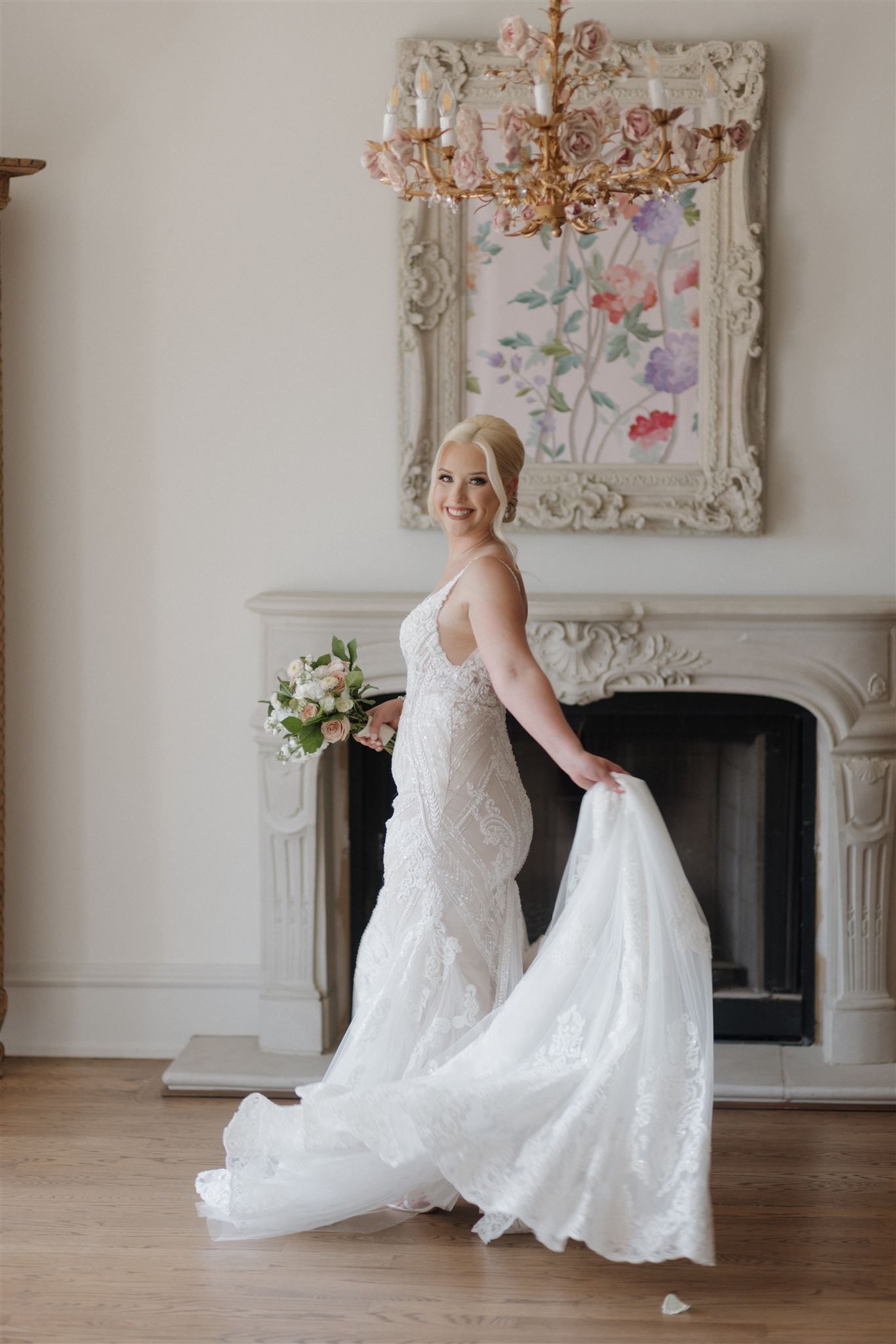 Wedding dress elegantly displayed near a fireplace with floral artwork and crystal chandelier.