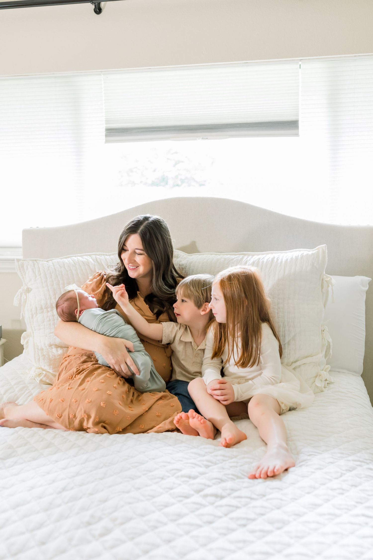 A family relaxes together on a white bed while holding a newborn baby.