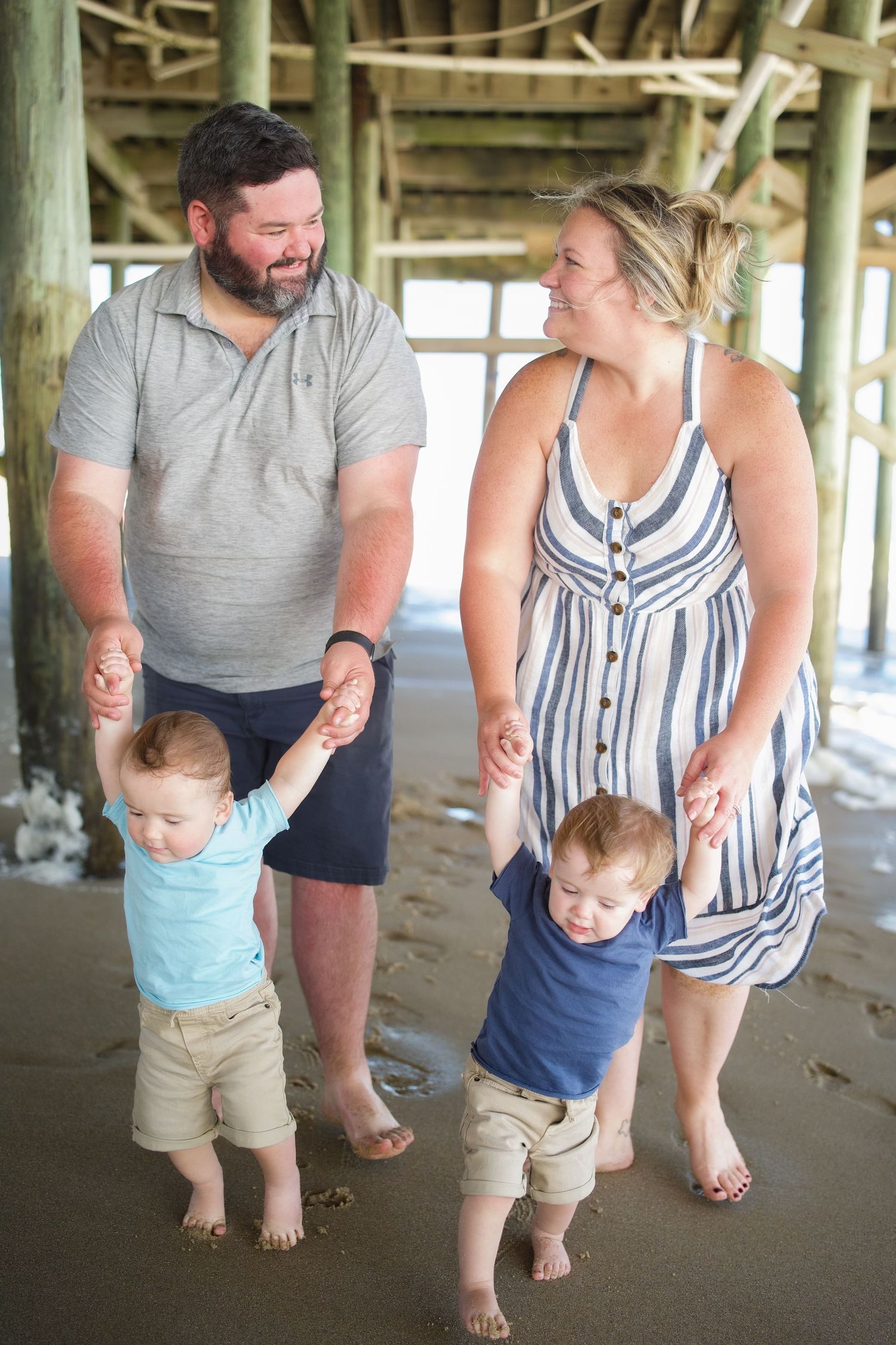 A family plays with their toddlers under a beach pier.