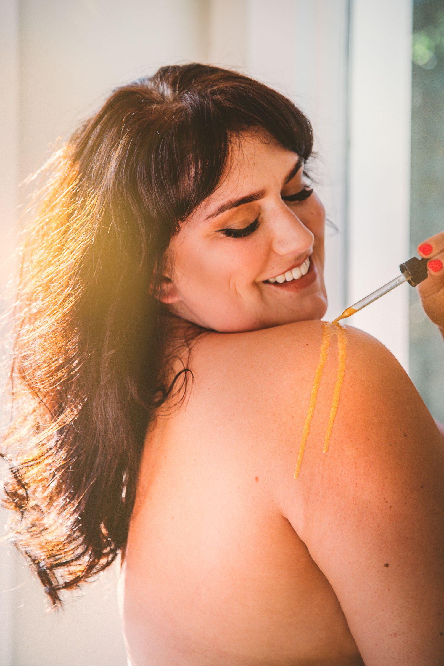 Brunette woman smiling down at shoulder, applying golden glow liquid