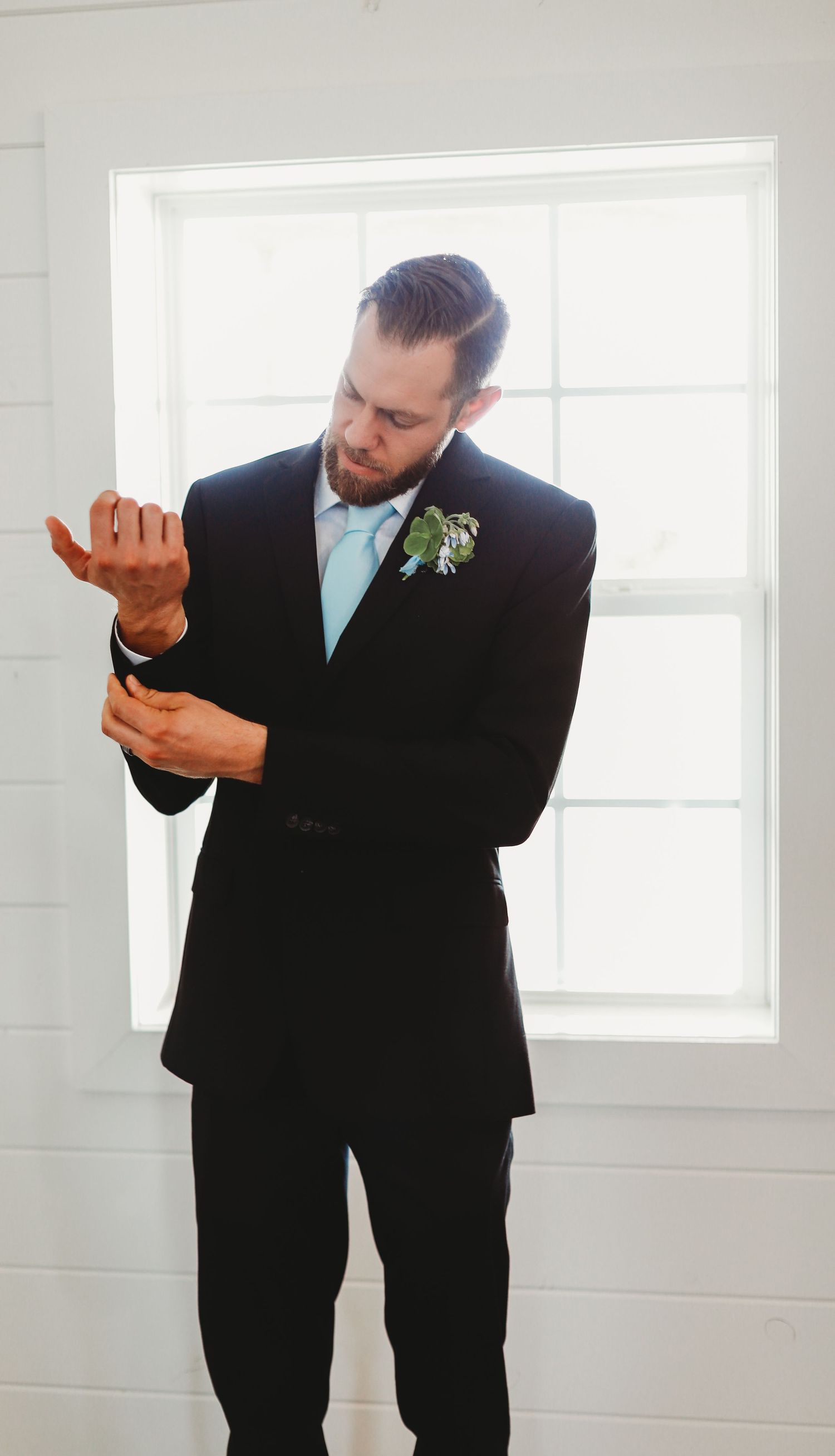 Groom in black suit and light blue tie adjusting cufflinks by window.