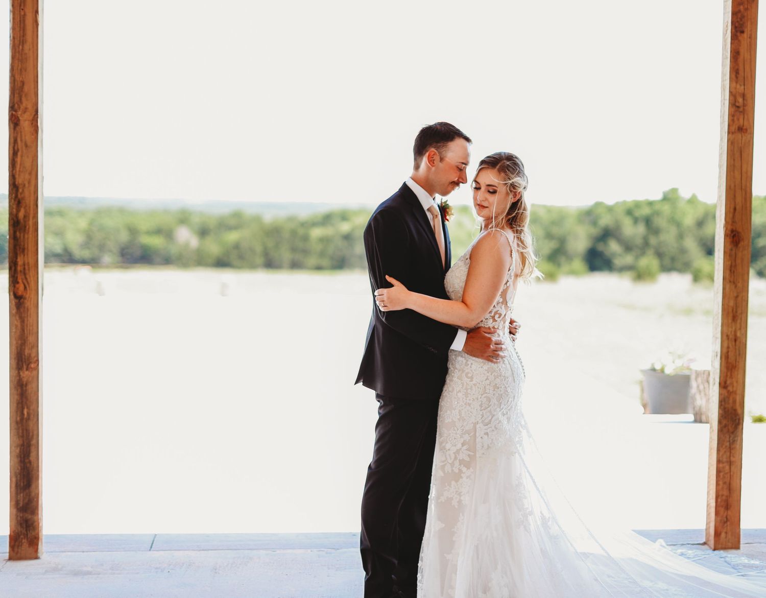 Wedding couple embraces in front of lakeside venue with white pillars.