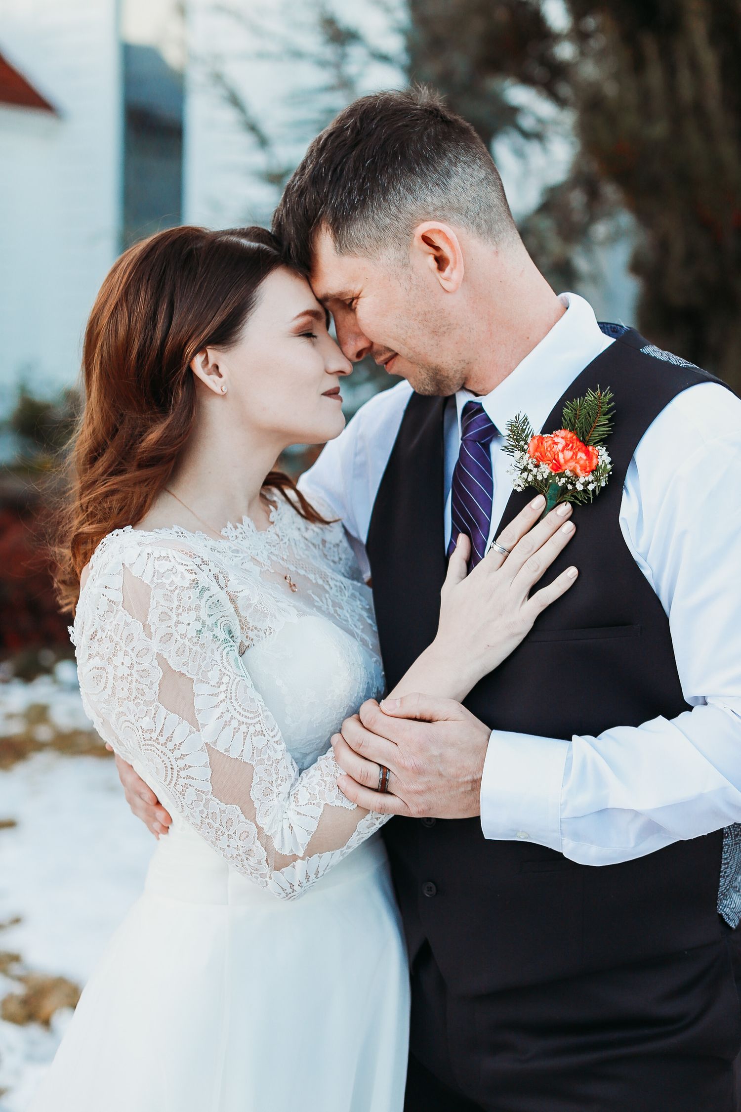 Wedding couple in lace dress and vest share a tender moment against a white building backdrop.