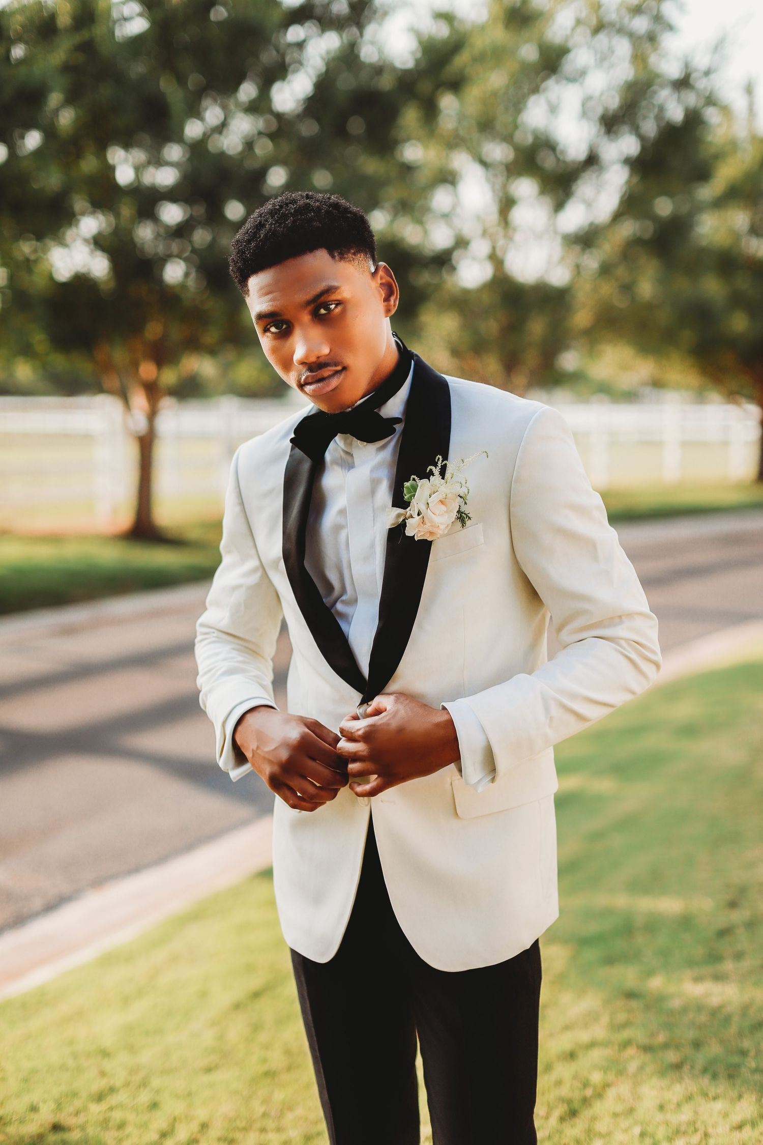 Elegant groom in white tuxedo jacket with black lapels and bow tie adjusts his cuffs outdoors.