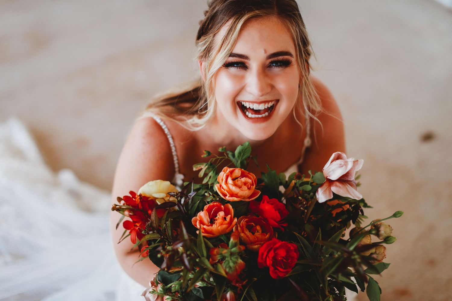 A bride laughs joyfully while holding a vibrant bouquet of red, orange and white roses during a wedding photoshoot.