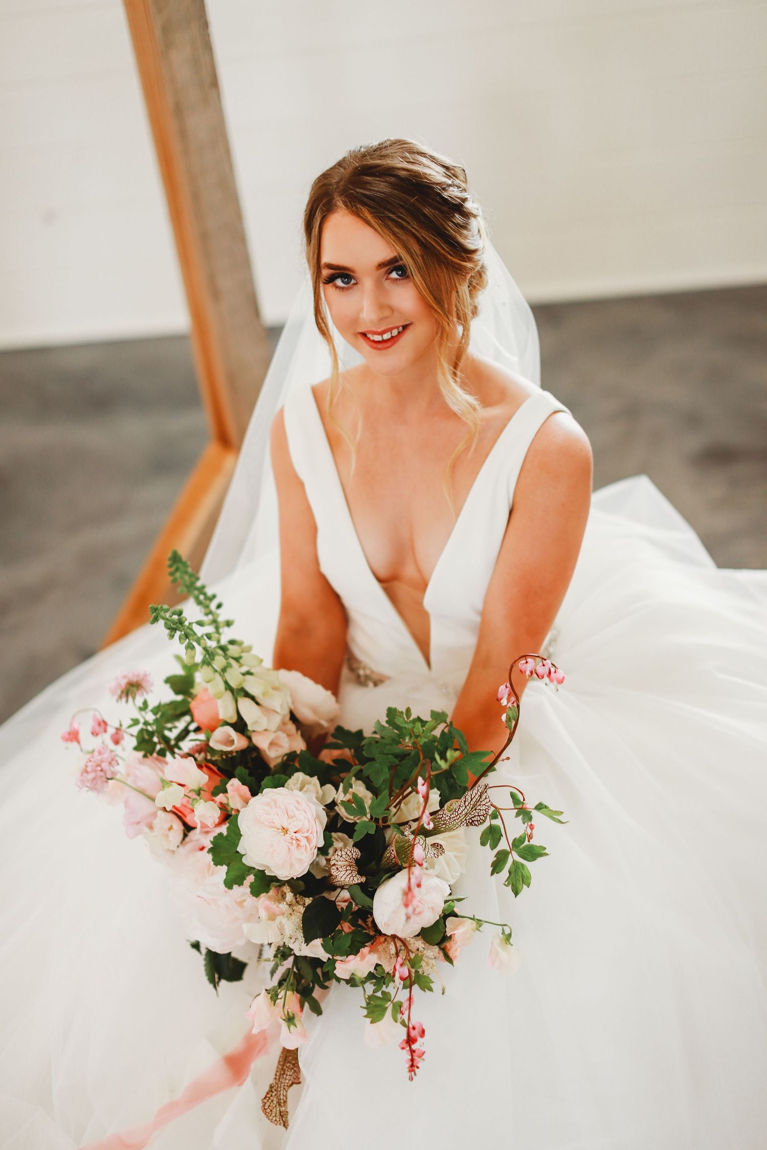 Bride in flowing white gown holding pink and peach floral bouquet under wooden arch.